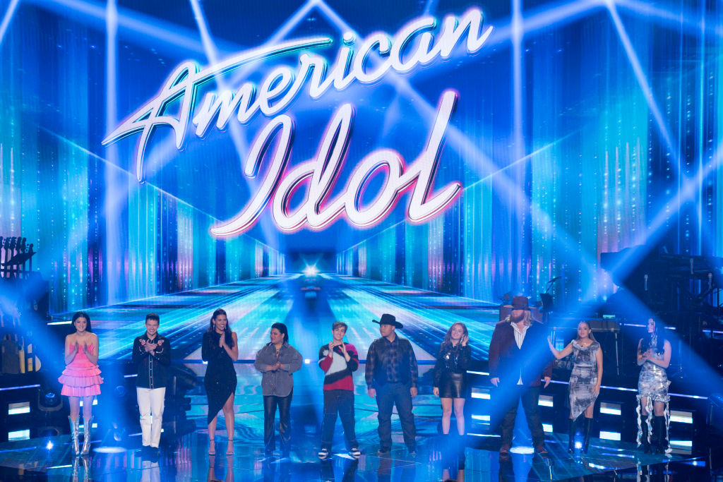 Contestants and judges of American Idol stand on stage under a large &quot;American Idol&quot; sign, dressed in various styles ranging from casual to formal