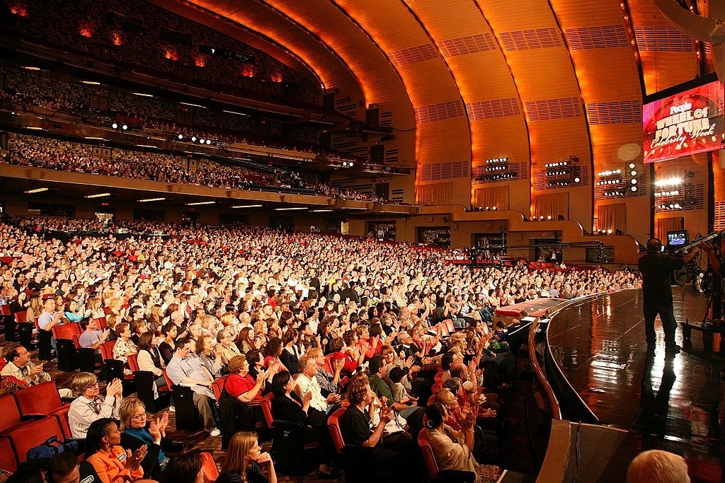 Packed audience in a grand theater, all seats occupied. Stage on the right with performers and crew members