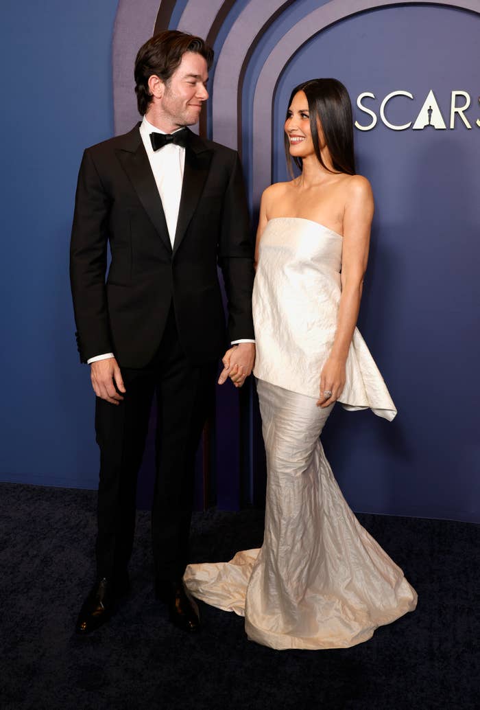 John Mulaney, in a classic tuxedo, and Olivia Munn, in an elegant strapless gown, holding hands on a red carpet at an Oscars event