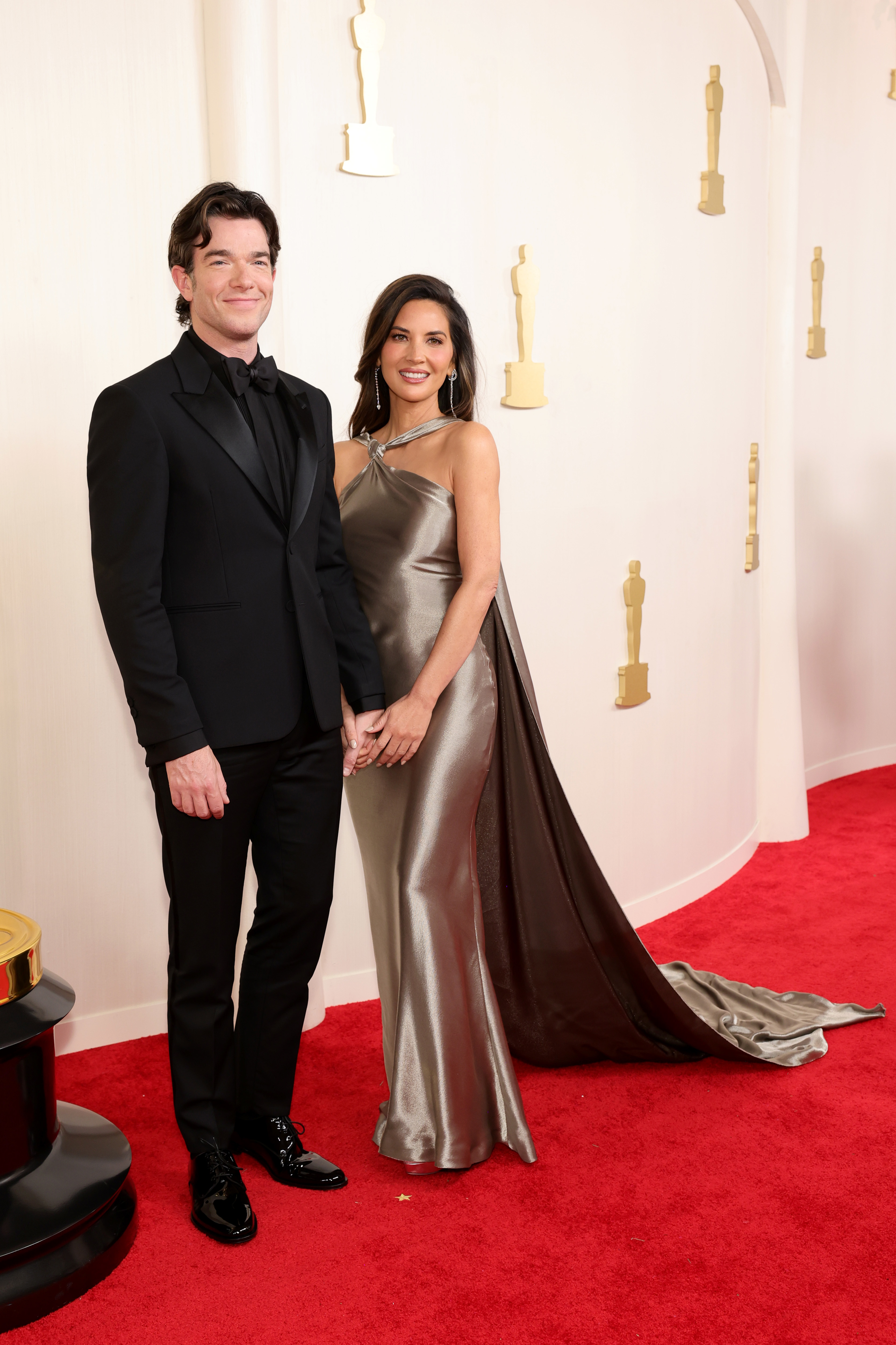 John Mulaney in a suit and Olivia Munn in a metallic gown pose on the red carpet at an event