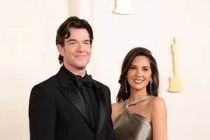 John Mulaney in a black suit and Olivia Munn in a metallic gown pose on the red carpet at an event