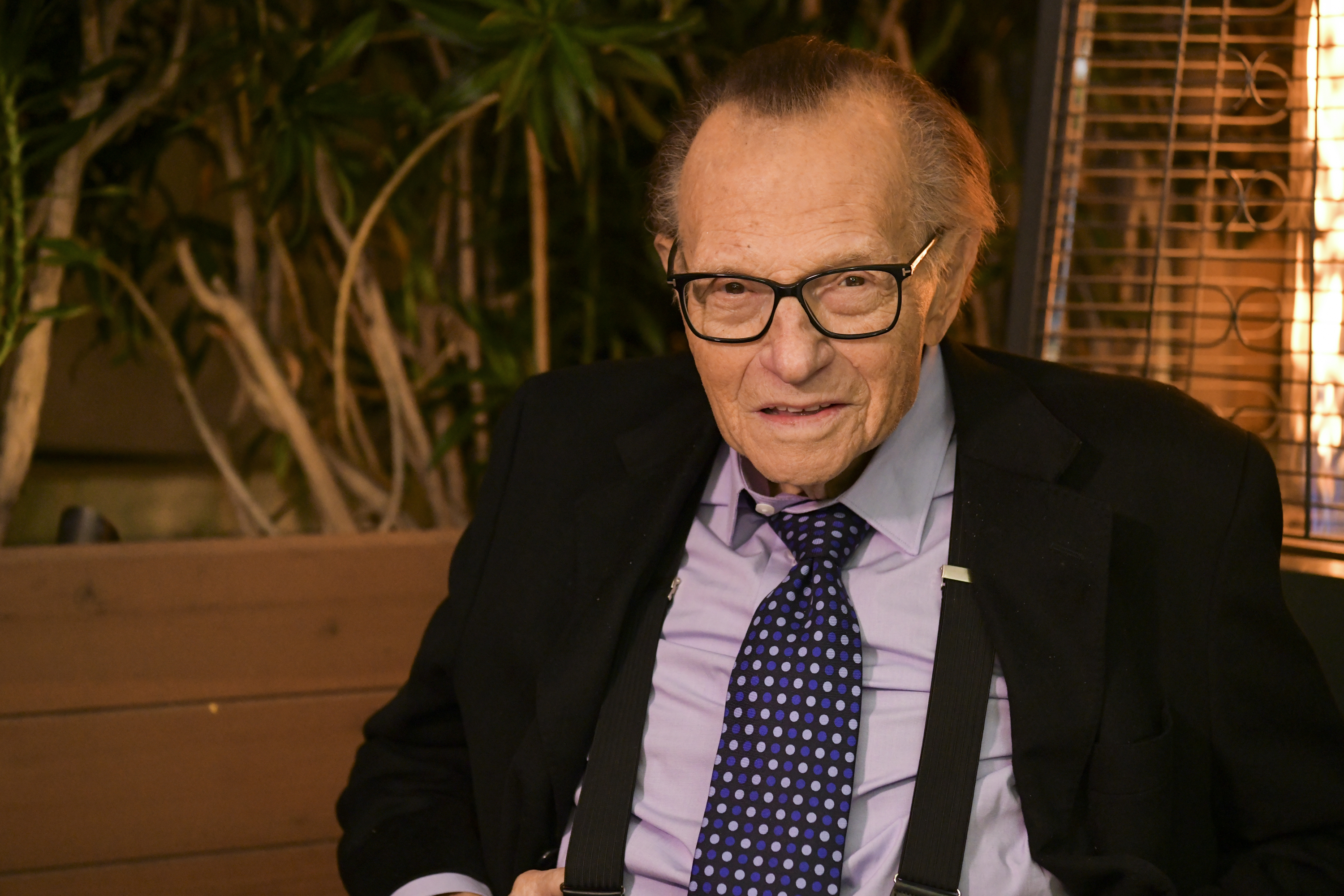 Larry King, wearing a suit and tie with suspenders, smiling while seated outdoors