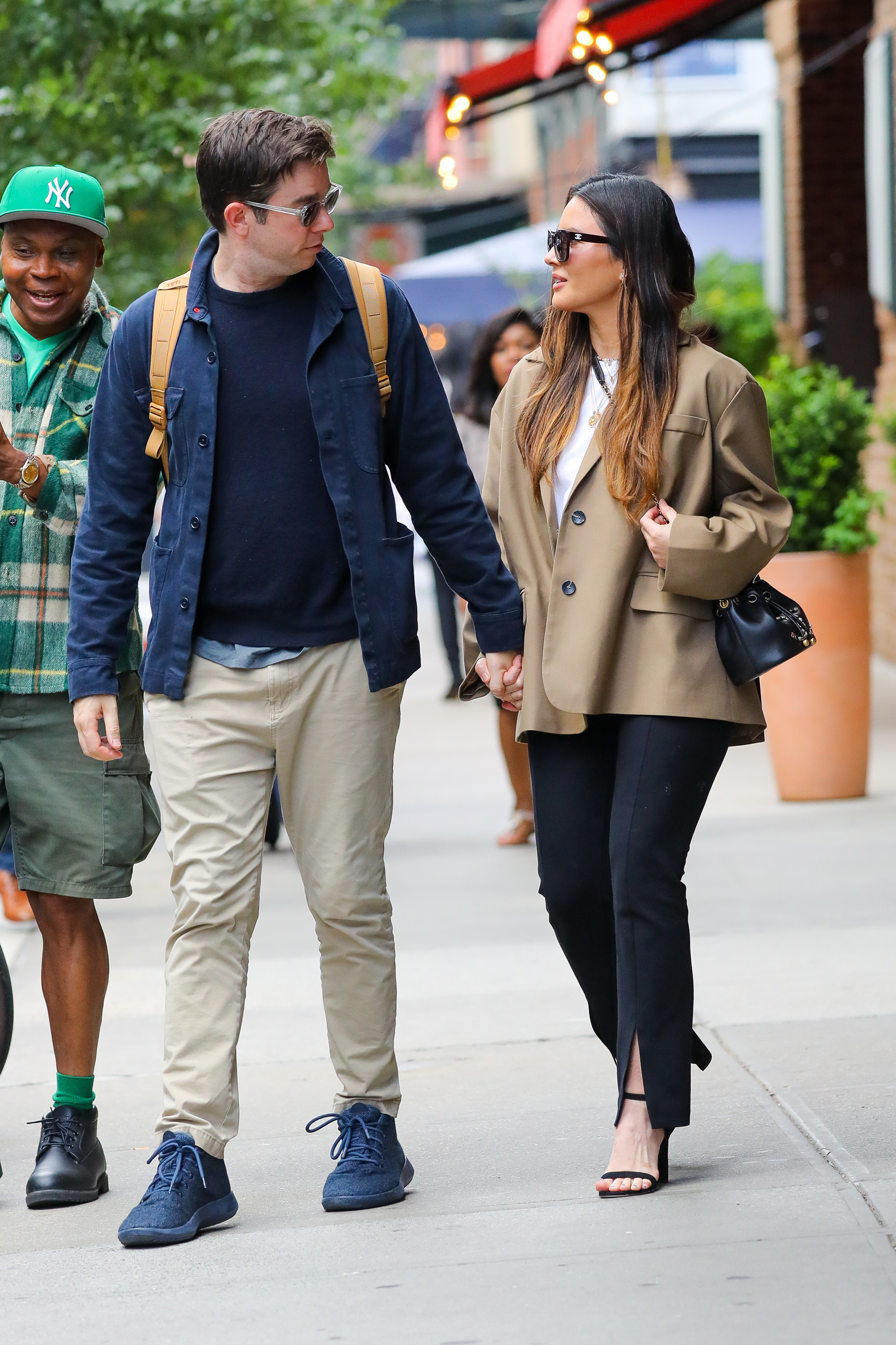 John Mulaney and Olivia Munn walk hand-in-hand on a city street; John wears casual attire with a backpack, and Olivia sports a blazer and sunglasses