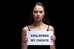 A woman holds a sign reading "Childfree by Choice." She has long hair and is wearing a sleeveless top