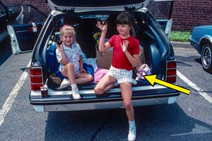 Two children sit in the open trunk of a car, each smiling and making a gesture. One girl is in a red shirt and shorts, while the other is in a striped outfit