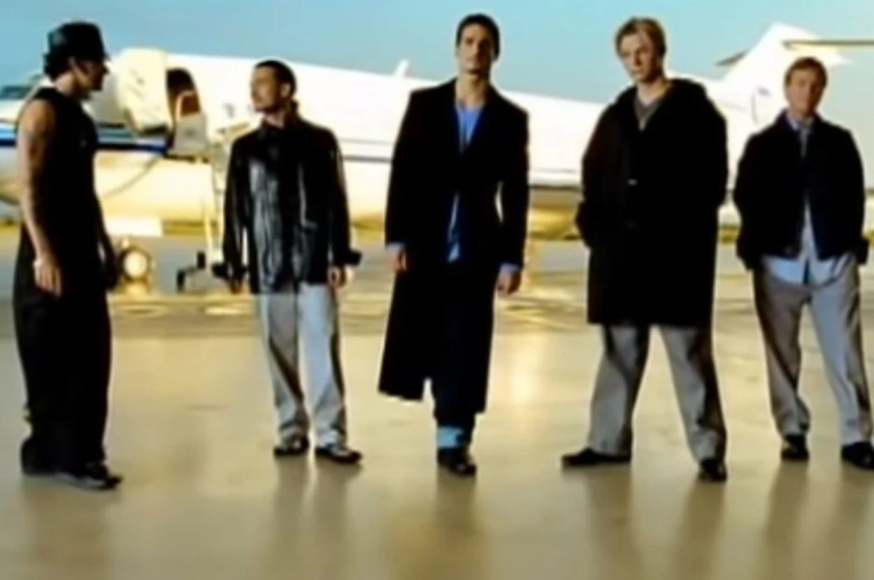 Five men stand in front of a private jet; one in a sleeveless shirt and hat, two in leather jackets, one in a long trench coat, and one in a casual coat and shirt