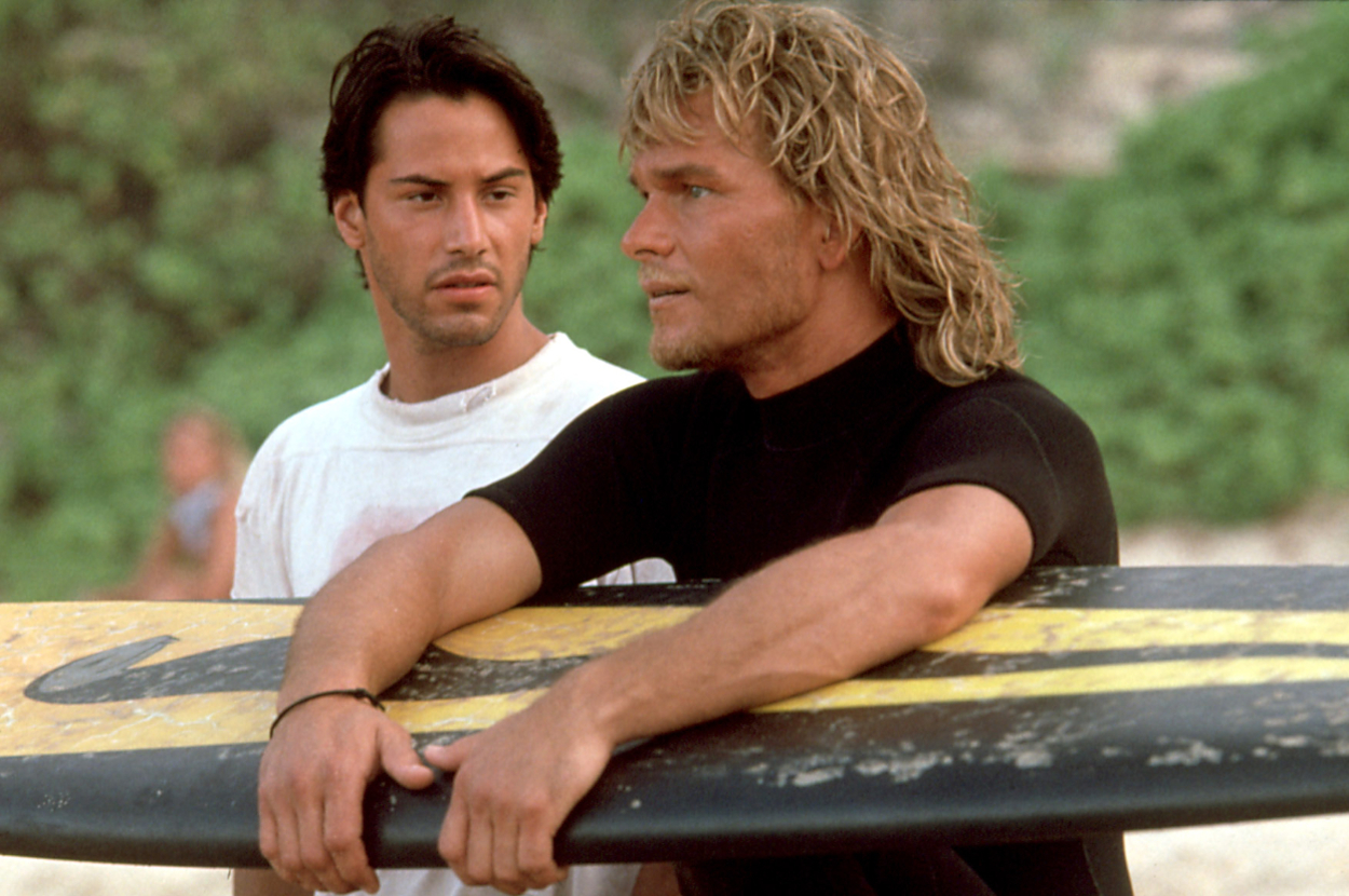 Keanu Reeves and Patrick Swayze in a beach scene from the movie "Point Break," with Patrick holding a surfboard and Keanu looking on