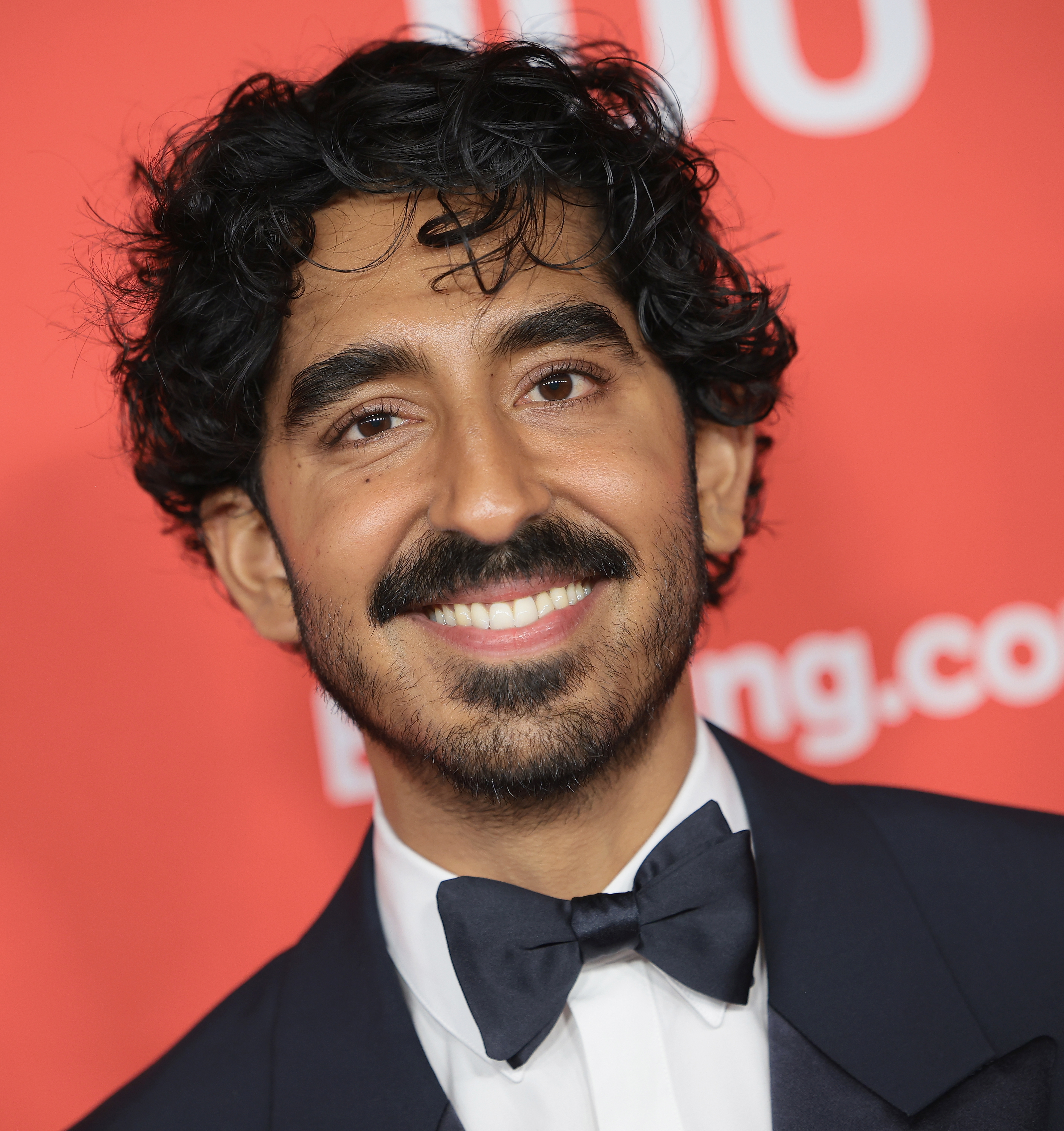 Dev Patel, smiling in a tuxedo with a bow tie, at a red carpet event