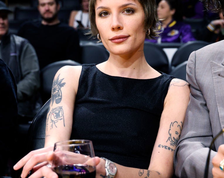 Halsey sits courtside at a basketball game, holding a drink and wearing a sleeveless top and jeans