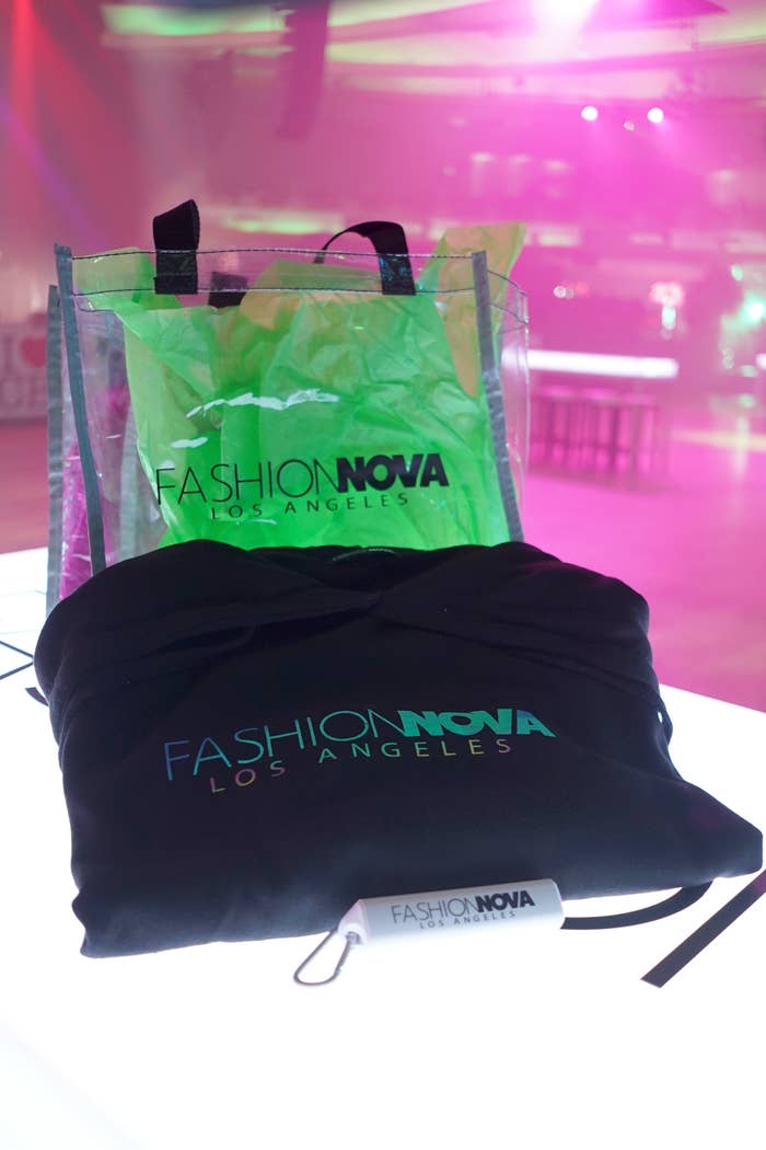 A Fashion Nova Los Angeles tote bag and drawstring bag are displayed on a table in a brightly lit room, with pink lighting in the background
