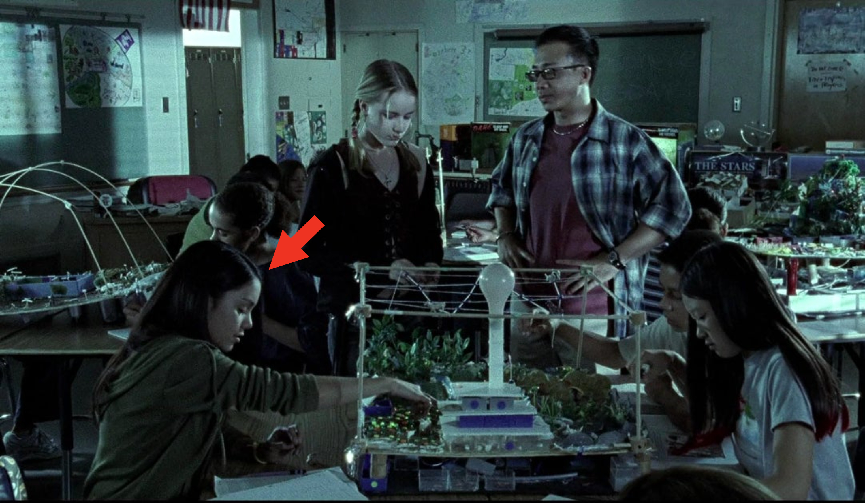 A classroom filled with students working on science projects. Two students, a girl and a boy, are standing next to a large model on a desk, discussing it