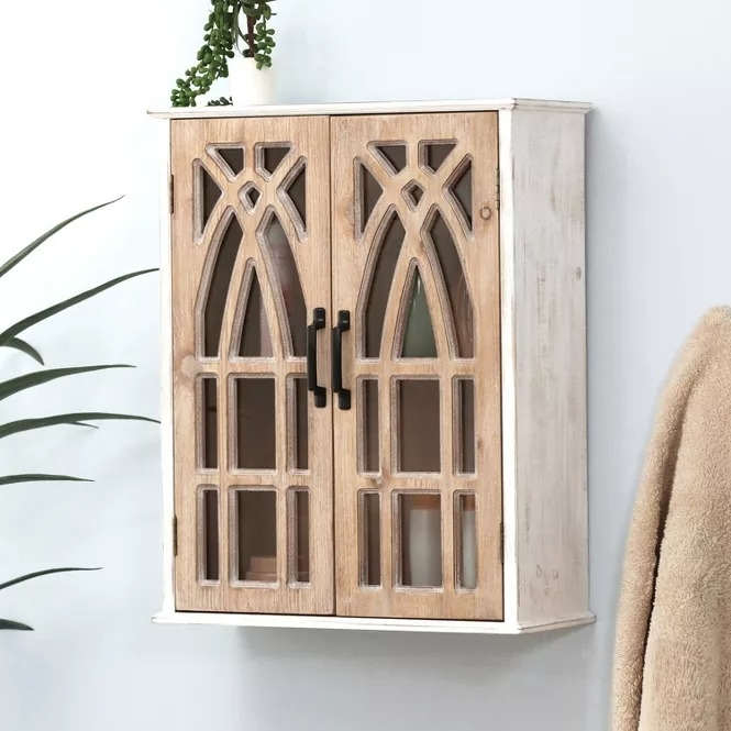 Wooden wall-mounted cabinet with decorative glass doors and black handles, featuring a small potted plant on top and part of a coat visible to the right