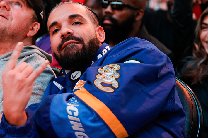 Drake sits by others wearing a blue jacket with his hand raised, gesturing a peace sign at a music event