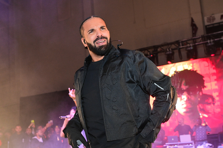 Drake performs on stage wearing a black jacket and gloves, with a backdrop and audience in the background