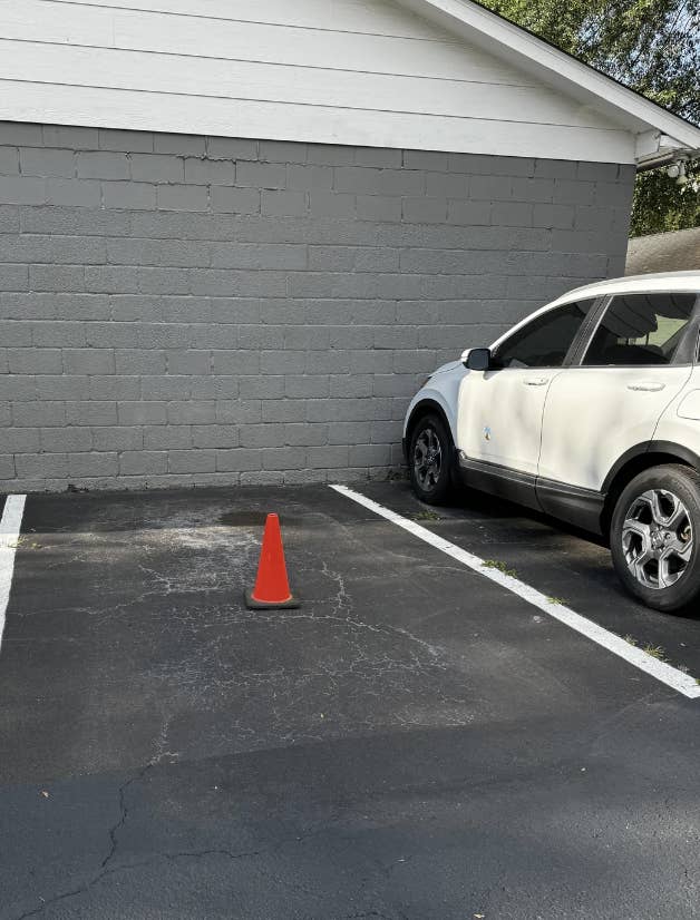 An empty parking space with a single orange cone in the middle, beside a white SUV parked next to a gray brick wall