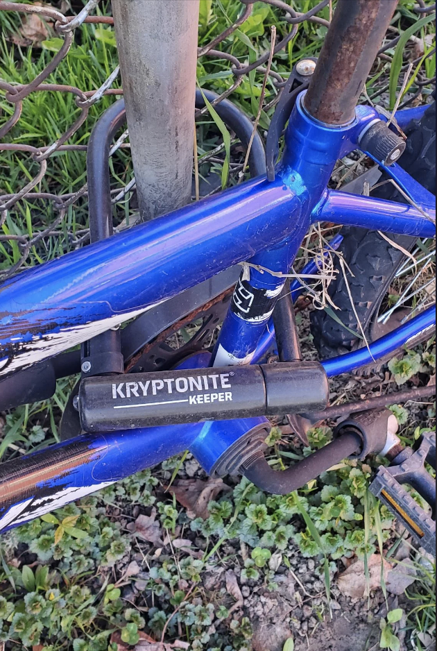 A blue bicycle secured to a pole with a Kryptonite Keeper bike lock. The bike is on a grassy and rocky surface