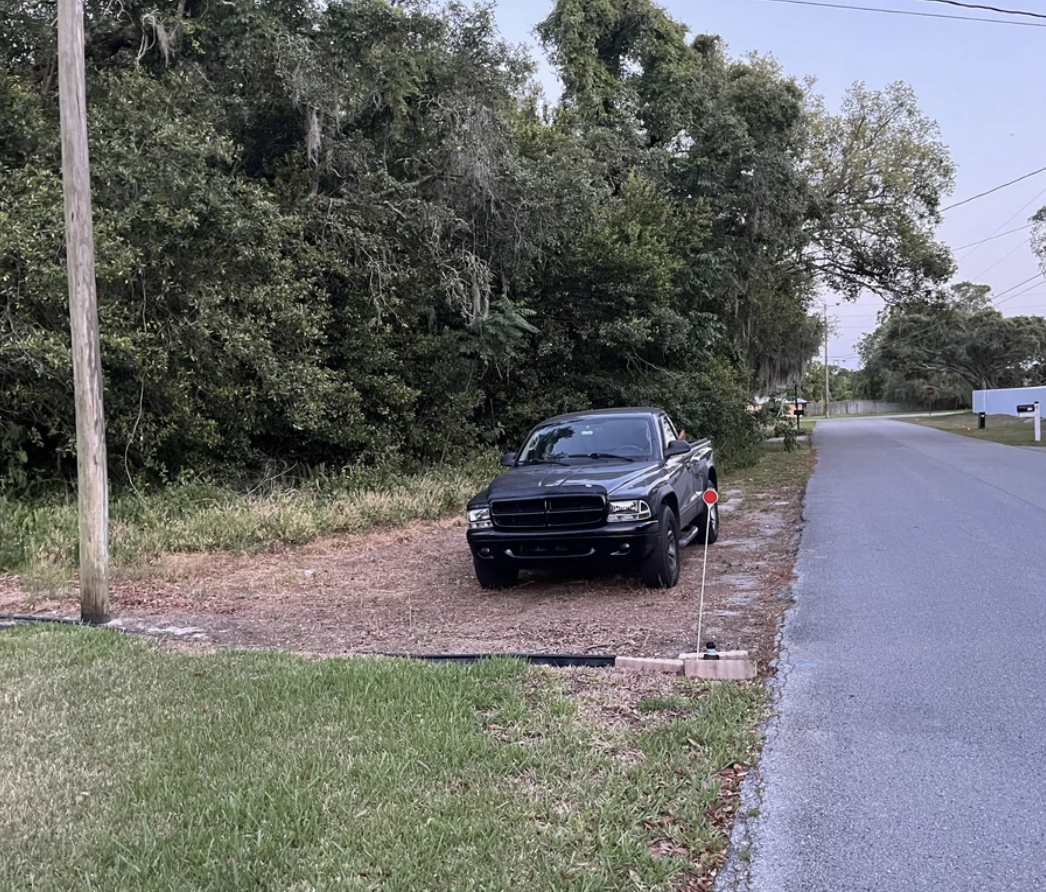 A black pickup truck is parked on a dirt patch next to a road, adjacent to a wooded area and utility pole