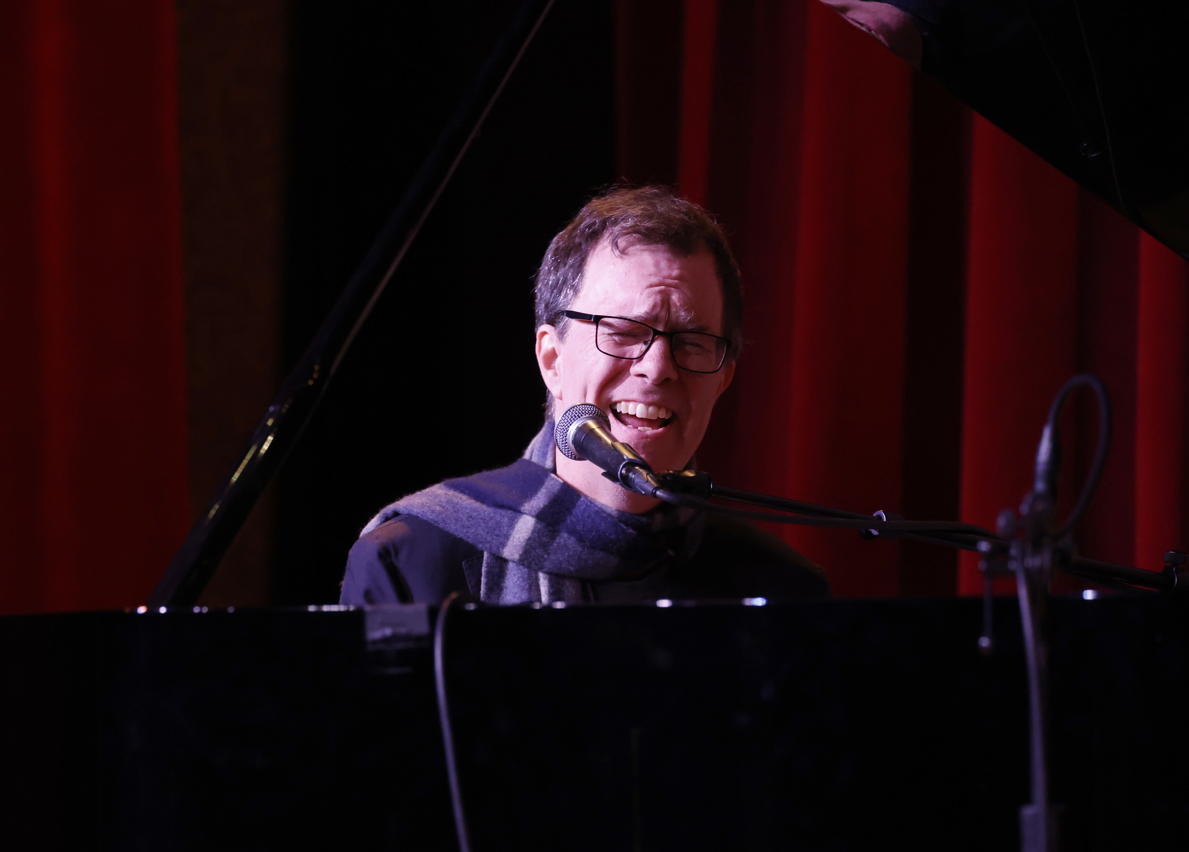 Ben Folds performs passionately on stage, playing the piano and singing into a microphone, dressed in a casual outfit with a scarf