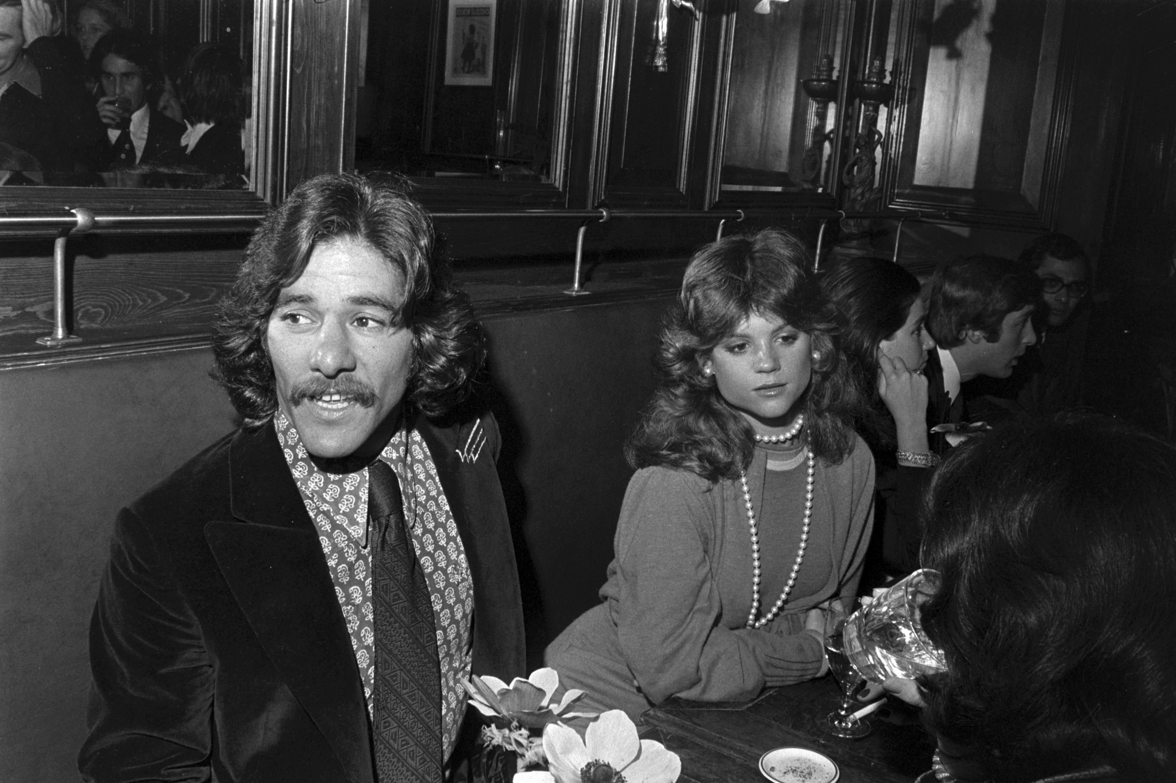 A man with a mustache and a woman wearing pearls sit at a table in a dimly lit venue with people in the background