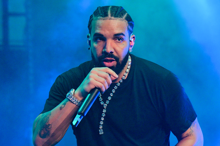 Drake performs on stage, wearing a black T-shirt, diamond necklace, and braided hairstyle, holding a microphone