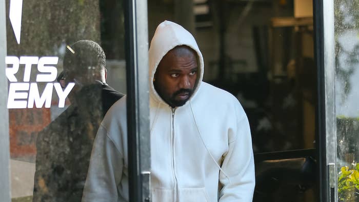 Kanye West is exiting a building wearing a white hoodie and matching pants. The reflection in the glass shows part of the &quot;Sports Academy&quot; sign