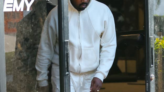 Kanye West is exiting a building wearing a white hoodie and matching pants. The reflection in the glass shows part of the "Sports Academy" sign