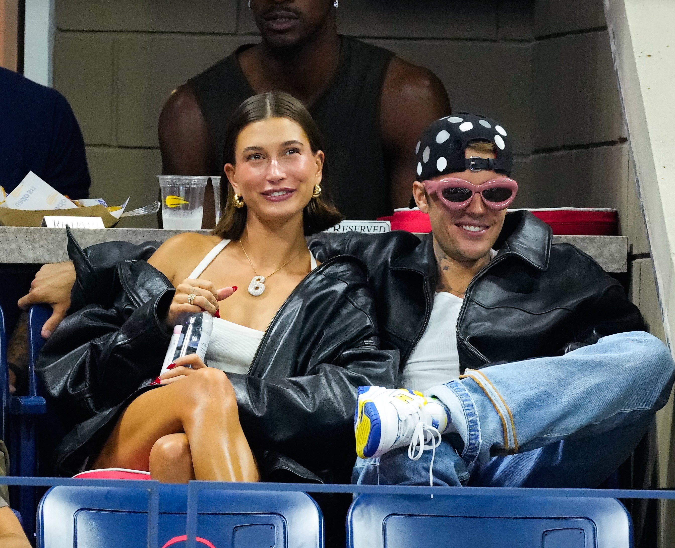 Hailey Bieber in a top and leather jacket with Justin Bieber who is wearing a dark jacket, sunglasses, and a spotted cap, seated at a sports event