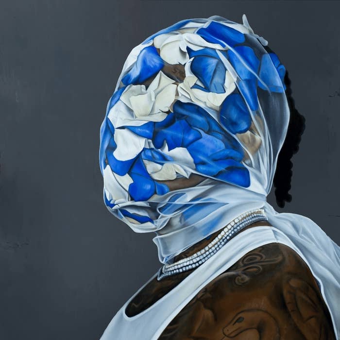A person is wearing a sheer veil covered in blue and white petals, obscuring their face entirely. They have visible tattoos on their shoulder and wear pearl necklaces