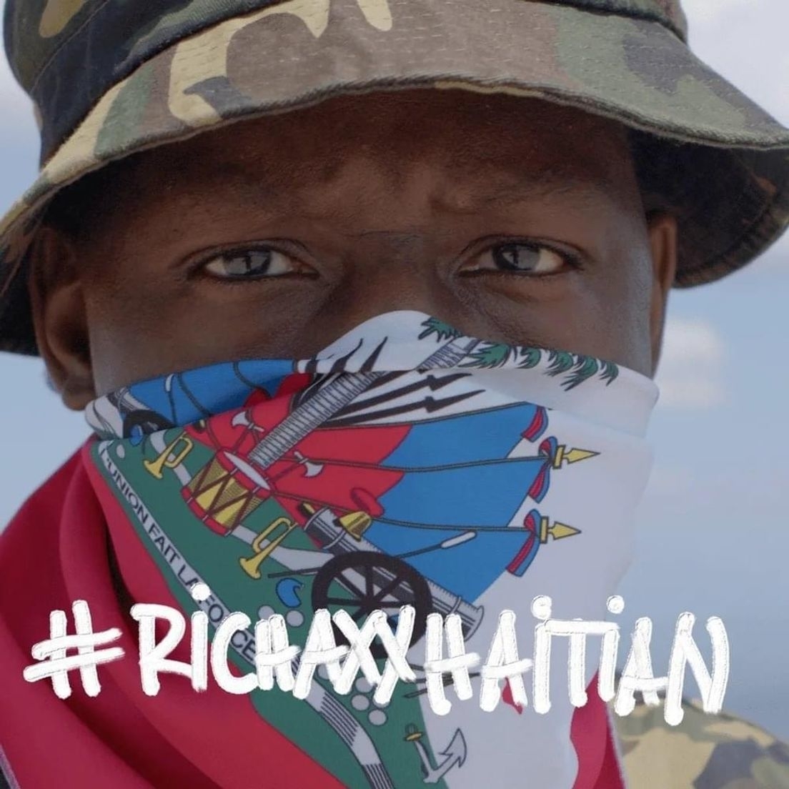 Man wearing a camouflage hat and a bandana with the Haitian flag design over his face. Text overlay says &quot;#RICHAXXHAITIAN.&quot;