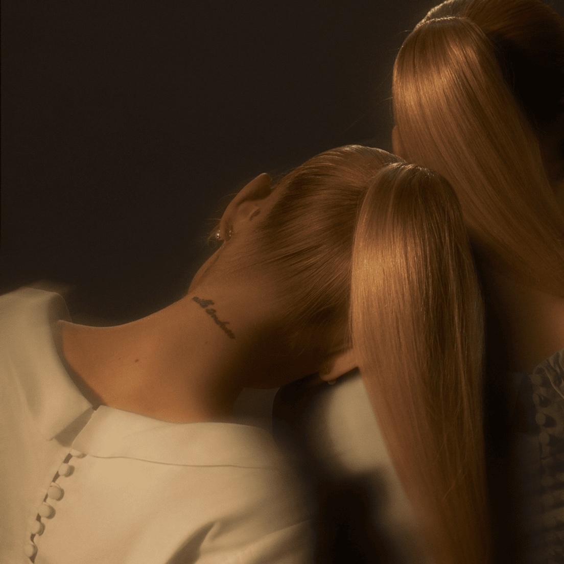 Two individuals with long, smooth hair tied back in ponytails, one person has a tattoo on the back of their neck. Their clothing style is minimalistic and formal