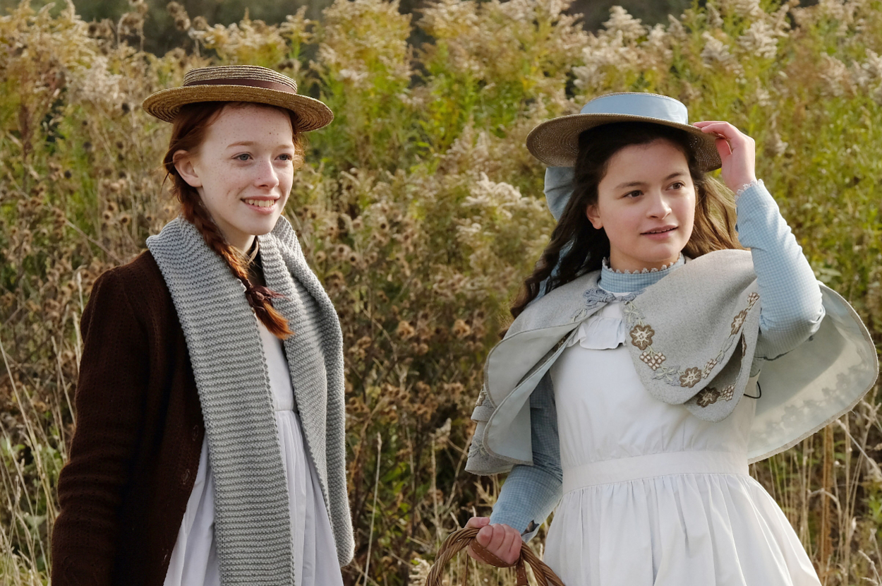 Anne (with braided hair and a straw hat) and Diana (in a bonnet with a cape) stand outdoors in a field, smiling