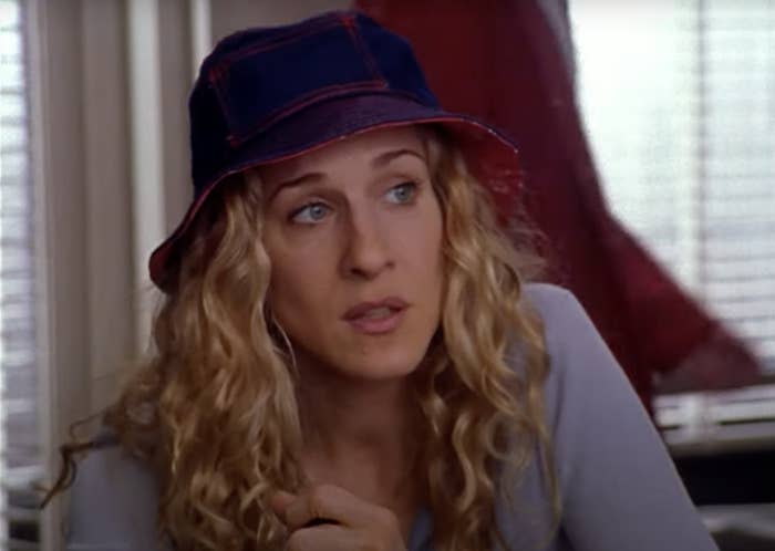 Sarah Jessica Parker, wearing a hat and with curly hair, is looking towards the side in a casual setting