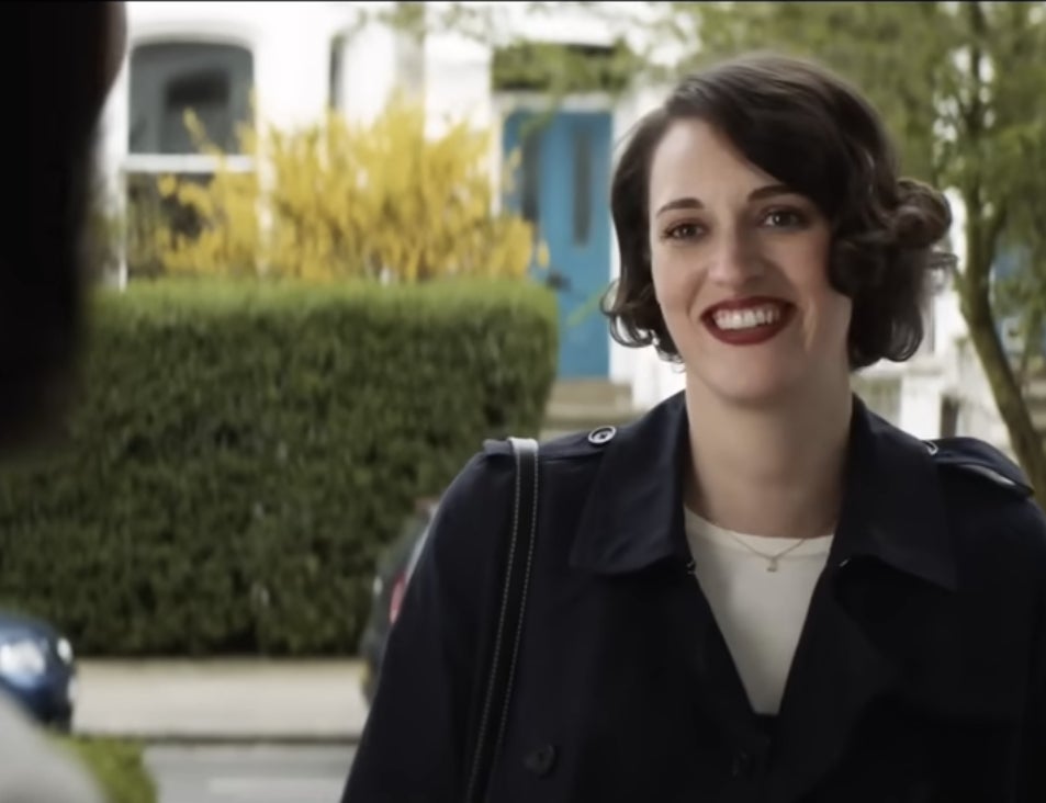 Phoebe Waller-Bridge smiles outdoors in a navy coat, standing in front of a building with a blue door