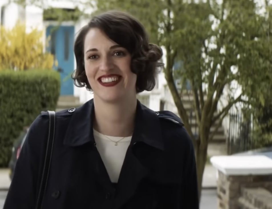 Phoebe Waller-Bridge smiles outdoors in a navy coat, standing in front of a building with a blue door