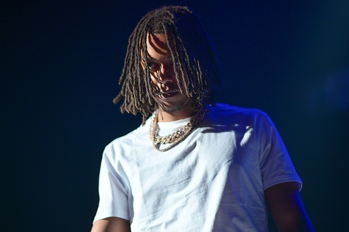 A person with shoulder-length hair wearing a white t-shirt and multiple gold chains around their neck stands under a spotlight on stage