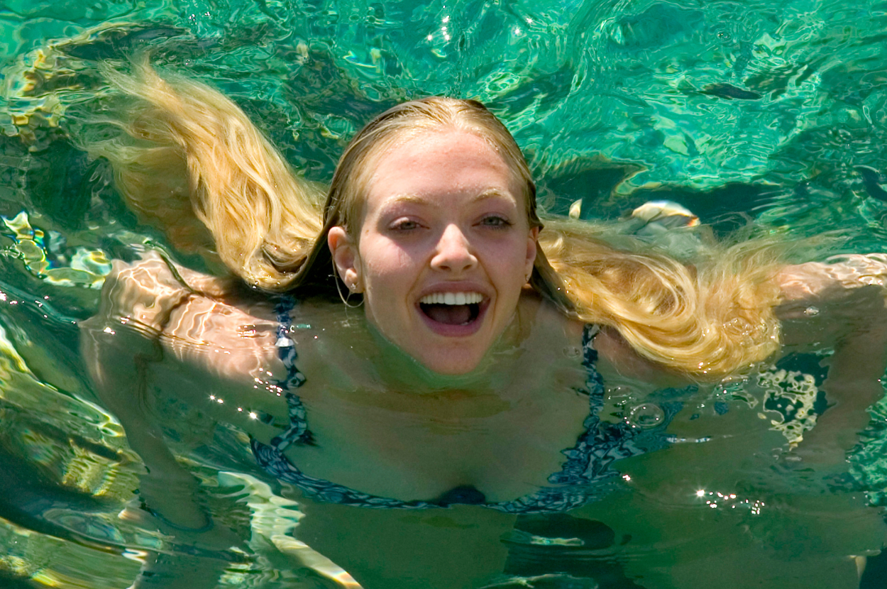Amanda Seyfried joyfully swims with her long hair fanned out in the water. She is wearing a patterned swimsuit and has an expression of happiness on her face