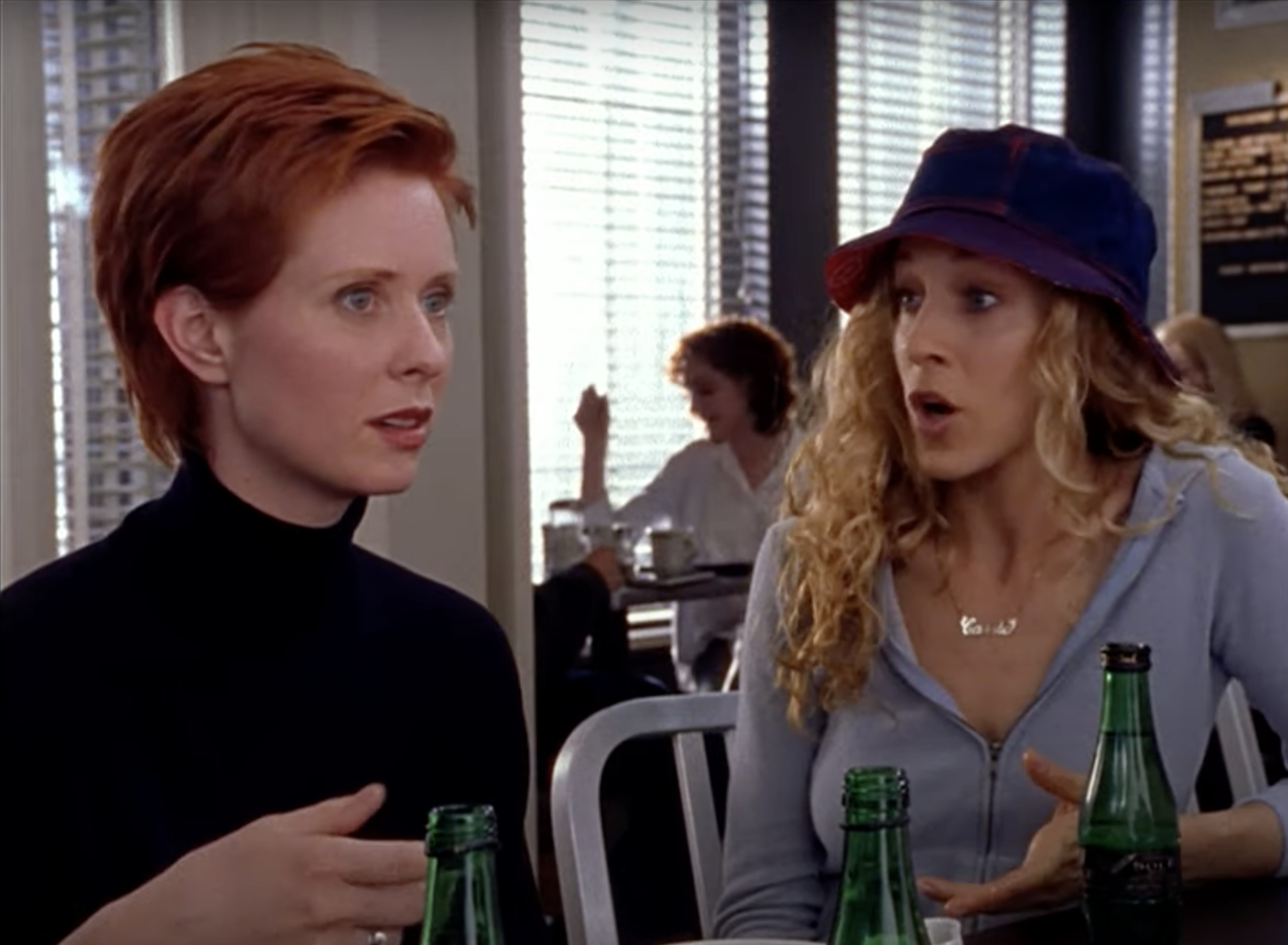 Cynthia Nixon and Sarah Jessica Parker are seated at a restaurant table, engaged in an animated conversation with bottles of sparkling water in front of them