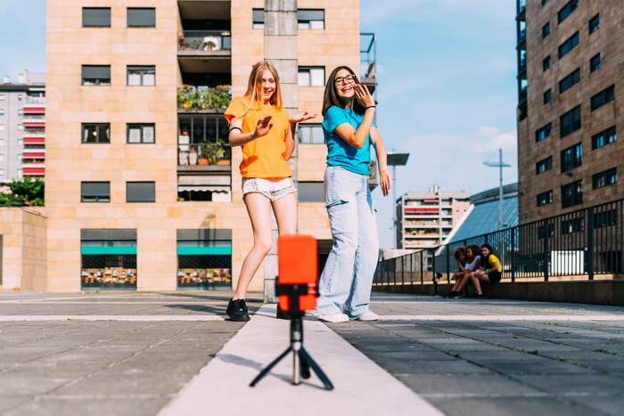 Two people, wearing casual clothes, are dancing in front of a smartphone on a tripod in an urban area with apartment buildings in the background