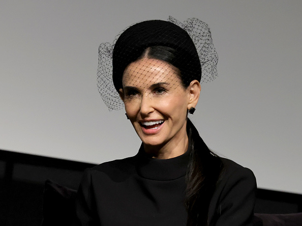 Demi Moore wearing a black dress with a black netted fascinator, smiling at an event