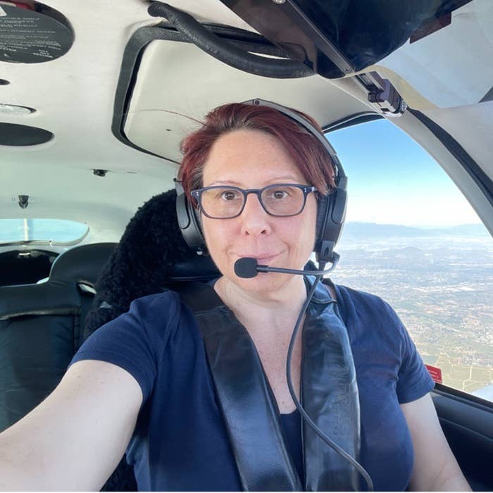 Teresa Polyak is seated inside an aircraft cockpit wearing a headset and glasses, with the blue sky and landscape visible through the window