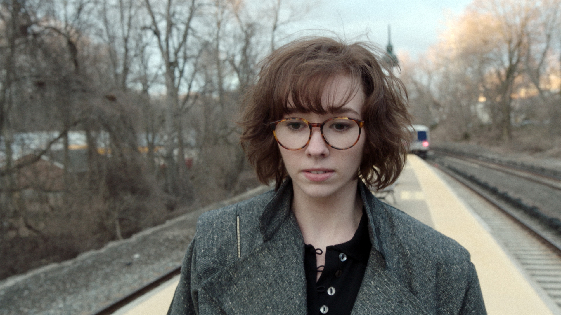 Woman with short, curly hair, wearing glasses and a gray coat, stands on a train platform with tracks and trees in the background
