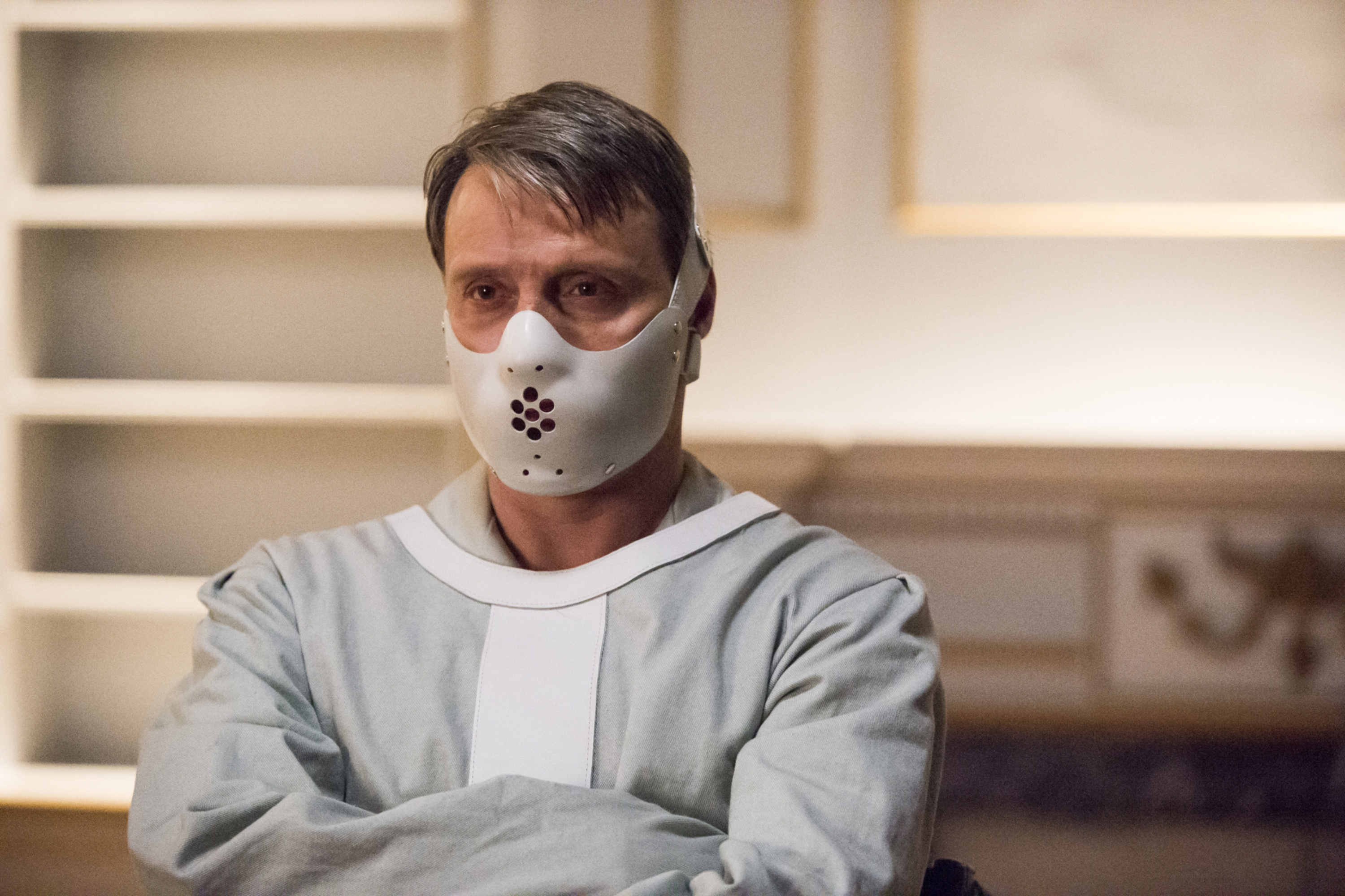 Mads Mikkelsen in a scene, wearing a white straightjacket and a mask covering his mouth, reminiscent of Hannibal Lecter
