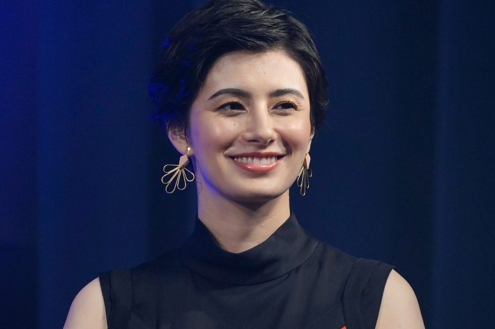 女性がカラフルな幾何学模様のドレスを着てマイクを持ちながら笑顔で立っています。