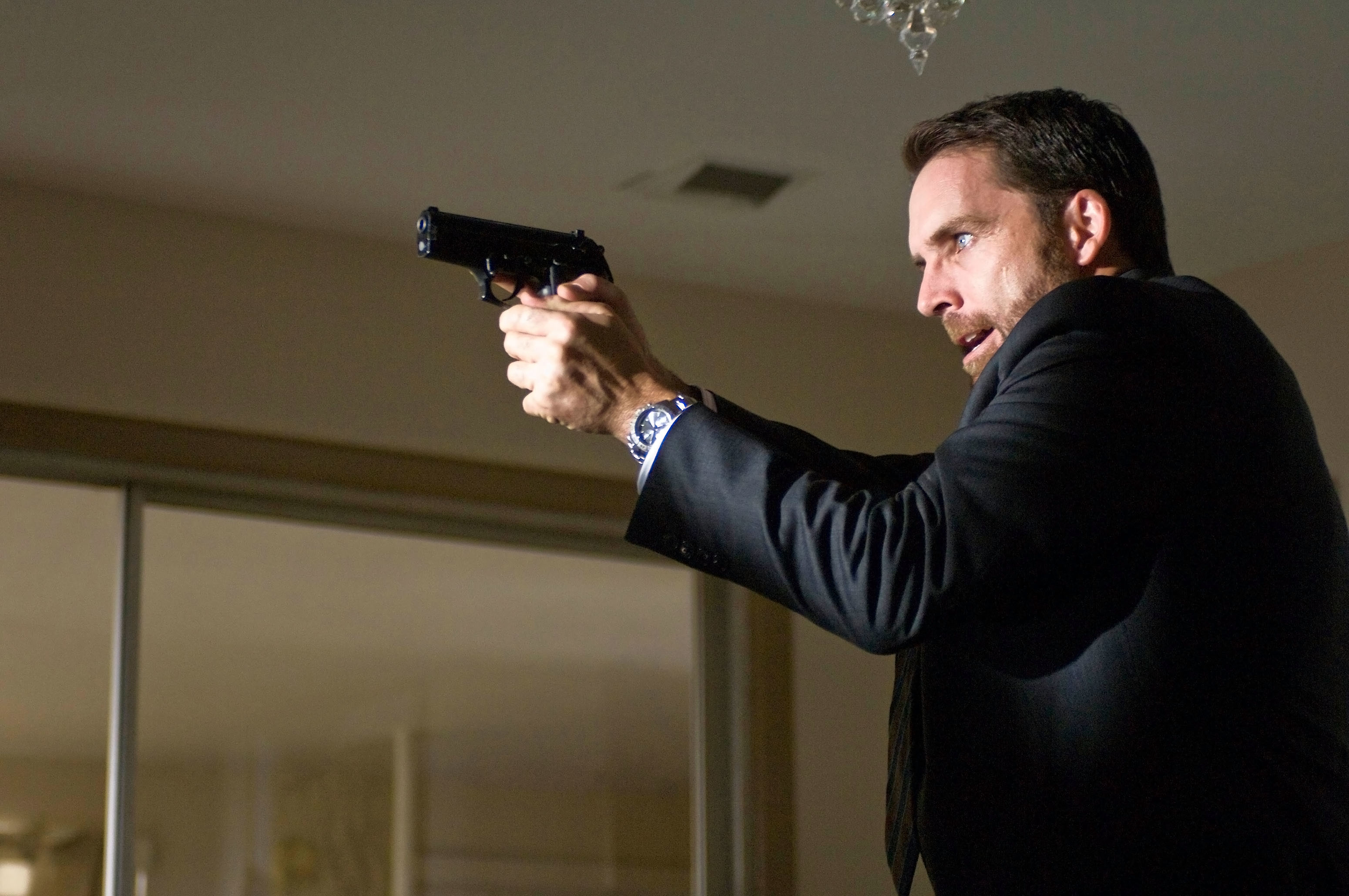 A man pointing a handgun, dressed in a suit, appears tense in an indoor scene
