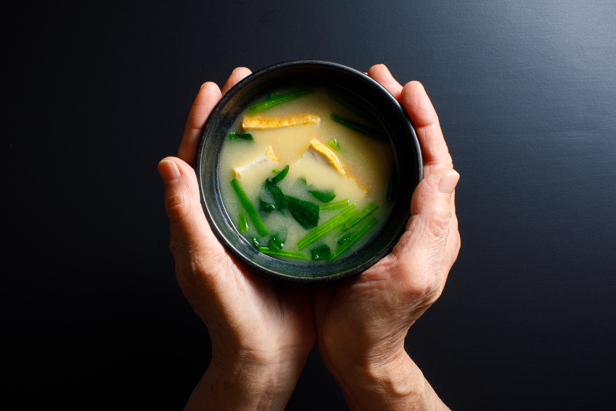 人の手に持たれたお椀の中に、味噌汁が入っている。味噌汁には豆腐、ネギ、ほうれん草が含まれている。