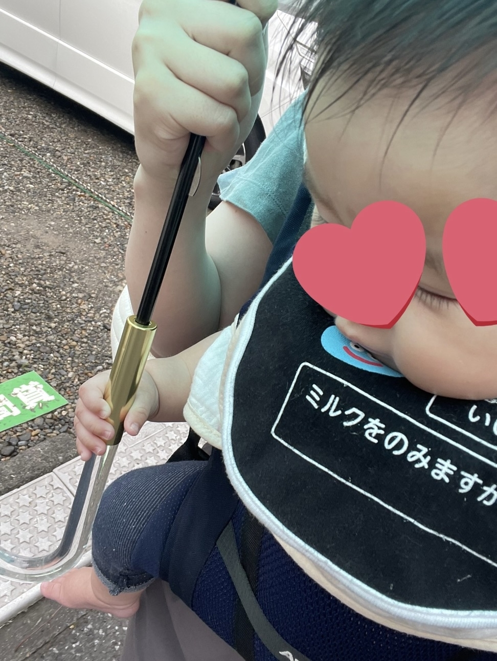 赤ちゃんが傘を持ちながら抱っこされている様子。顔の目の部分がハート型で隠されている。