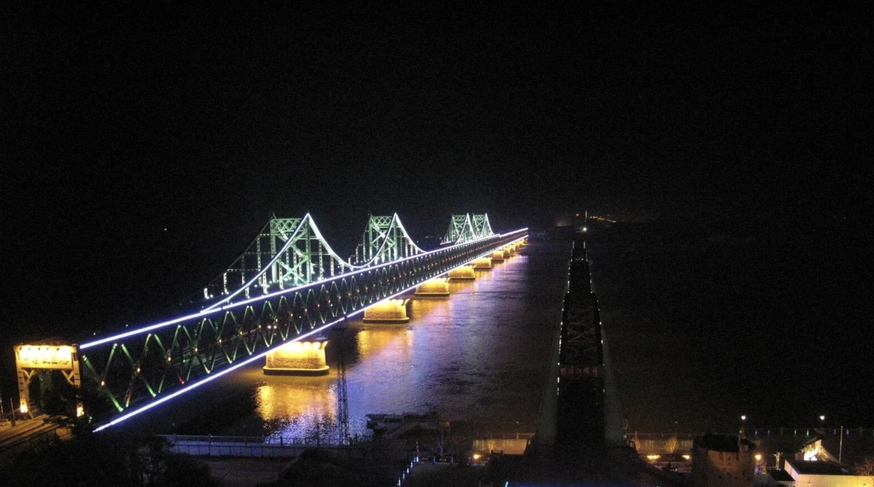 A night view of a brightly lit long bridge with multiple triangular supports spanning over a wide body of water, showcasing a modern architectural design
