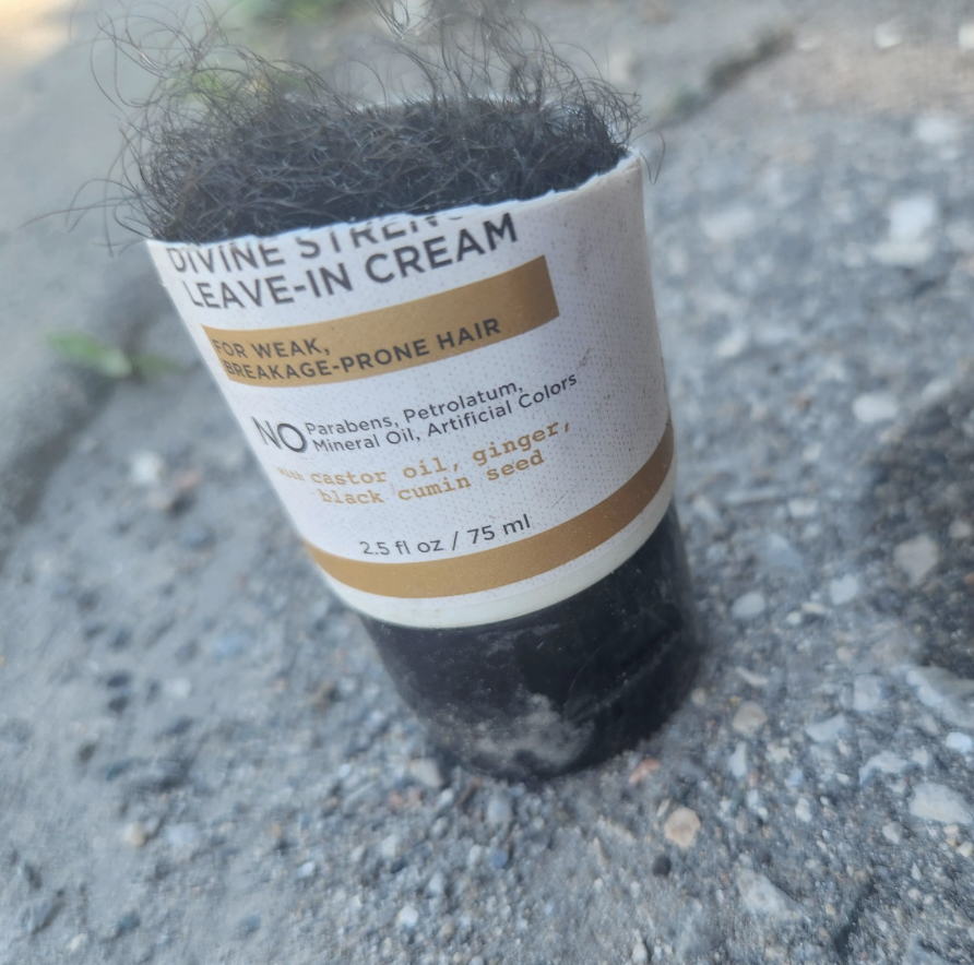 A 75mL container of Divine Strength Leave-In Cream for weak, breakage-prone hair is lying on the ground, covered in what looks like hair and debris