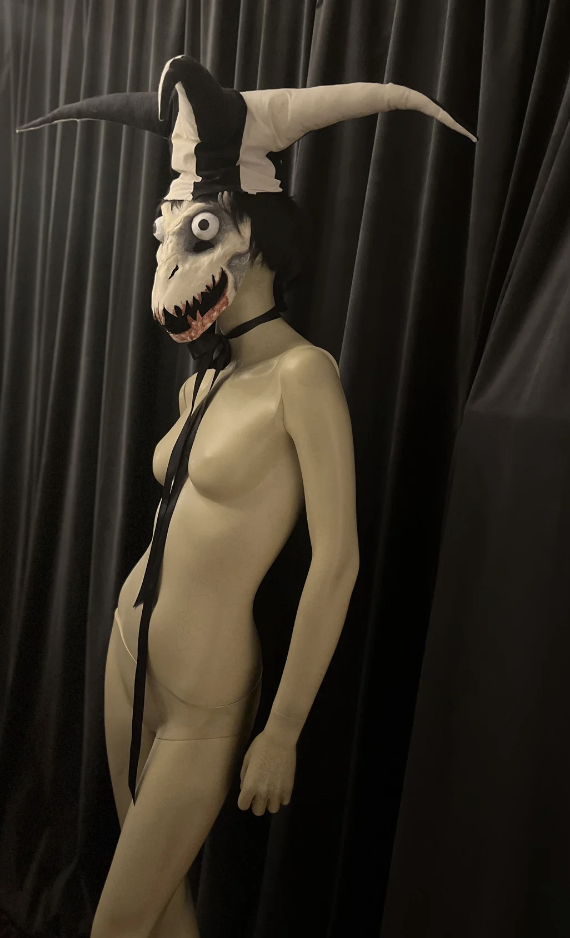 A mannequin dressed in a black and white jester hat with a scary mask, standing against a dark curtain background