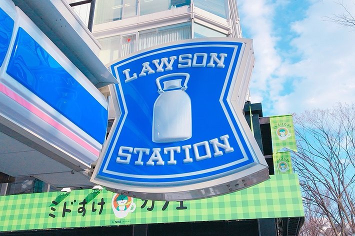 ローソンステーションのサインが映っている街の風景。看板には牛乳瓶のロゴ。ビルや木々が背景に見える。